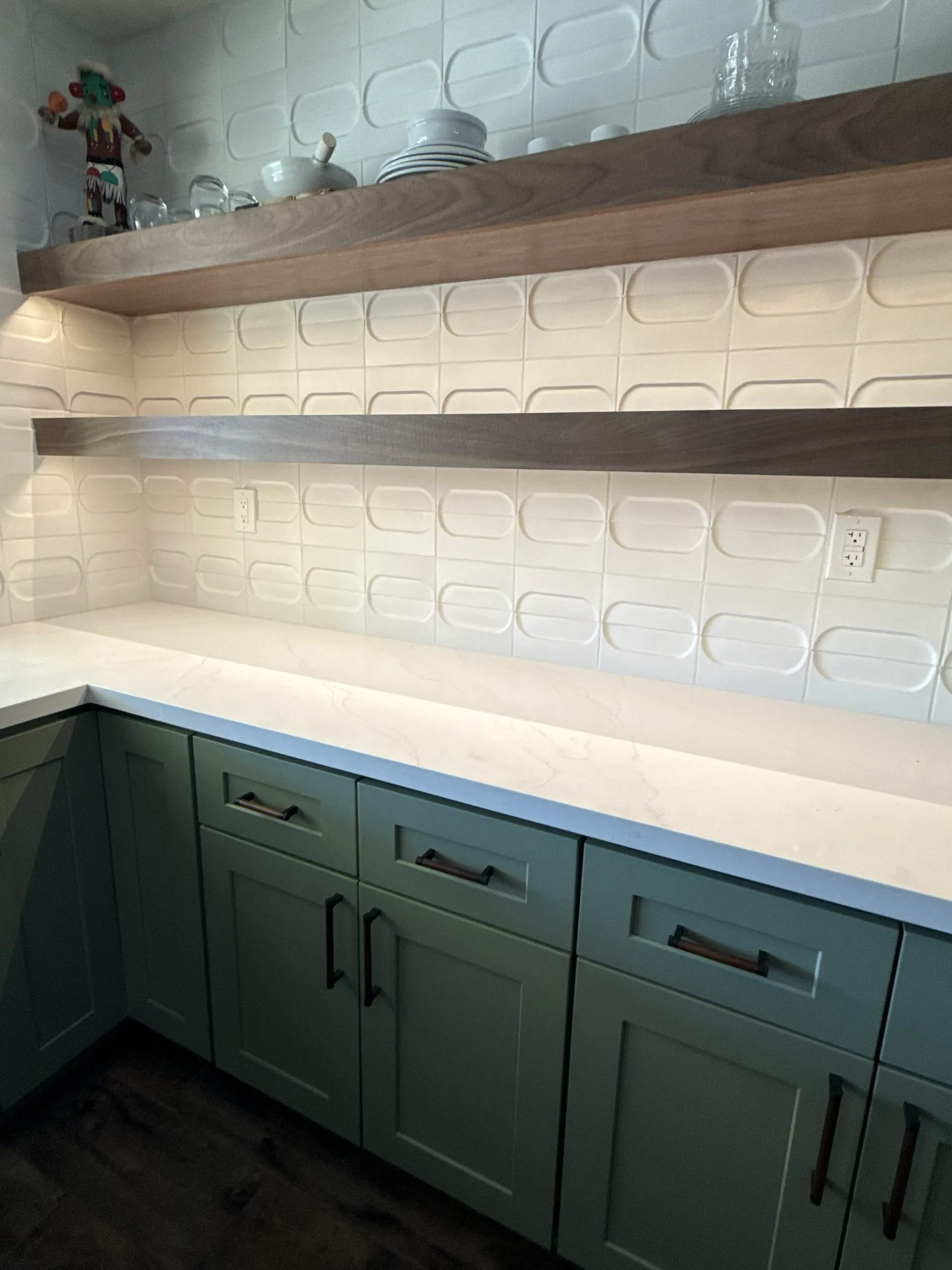 Detail of textured white wall tile with walnut floating shelves and green cabinets